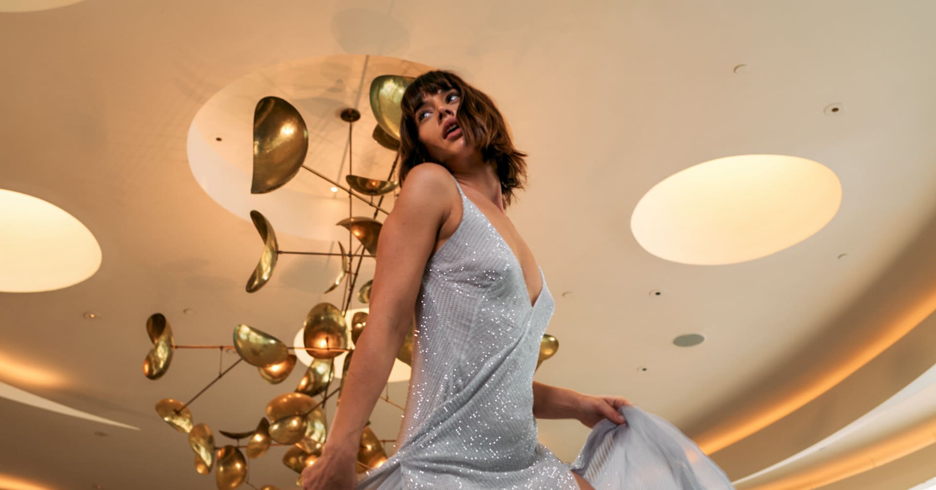 Person wearing a silver dress posing indoors beneath decorative ceiling lights.