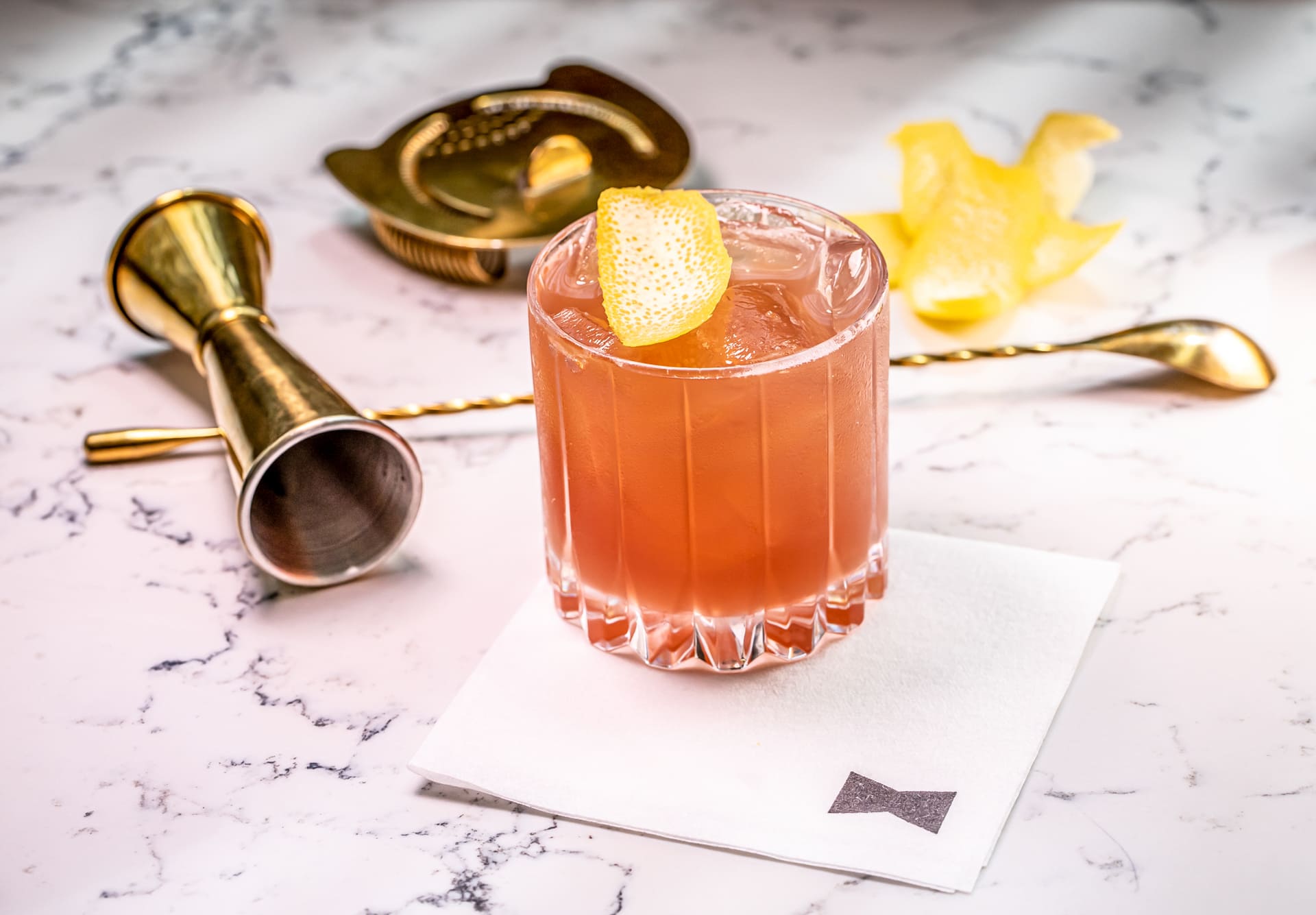 Pink cocktail with ice and lemon peel garnish on a bar surface with bar tools.