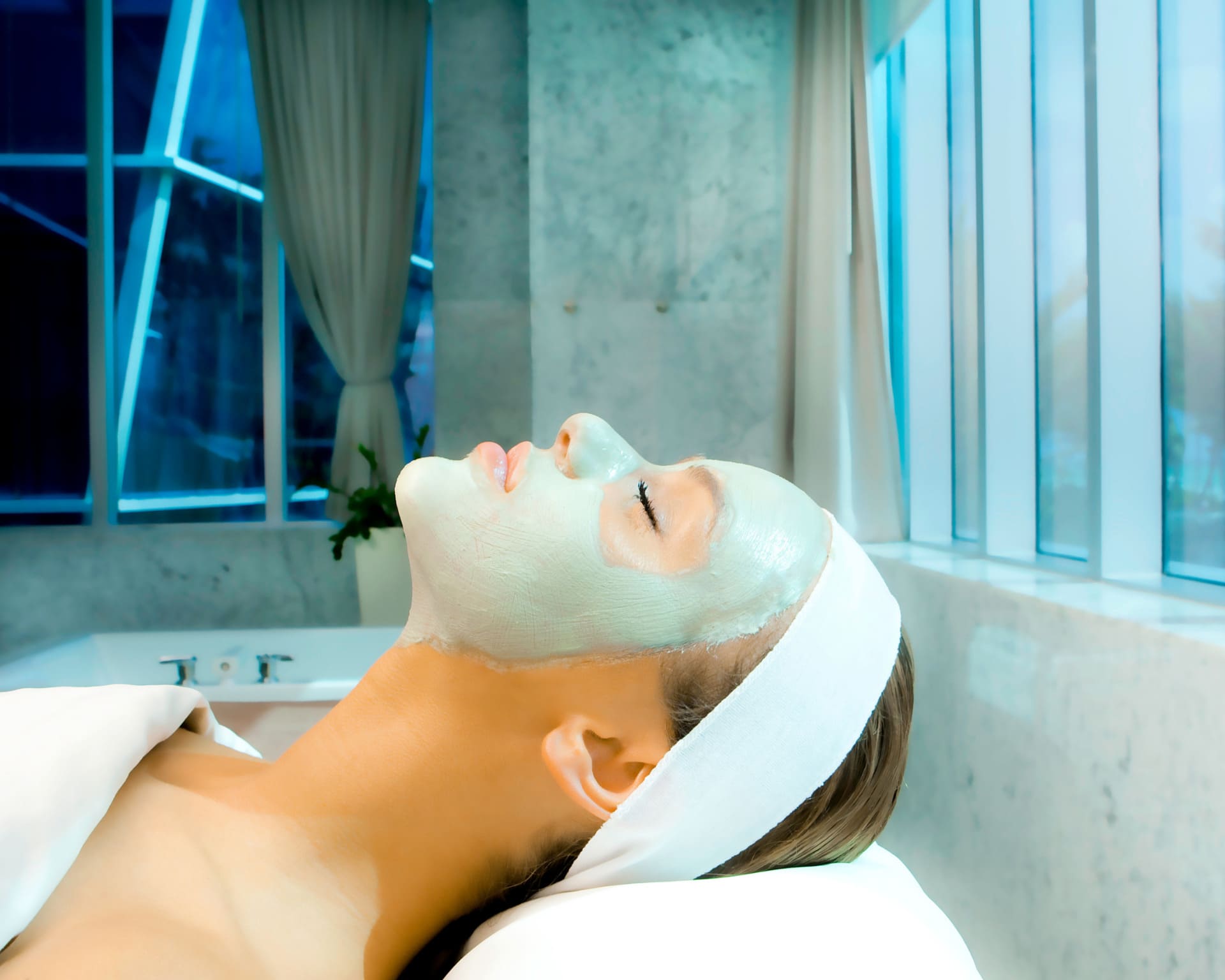 Person lying down with eyes closed while receiving a facial mask treatment in a spa room.