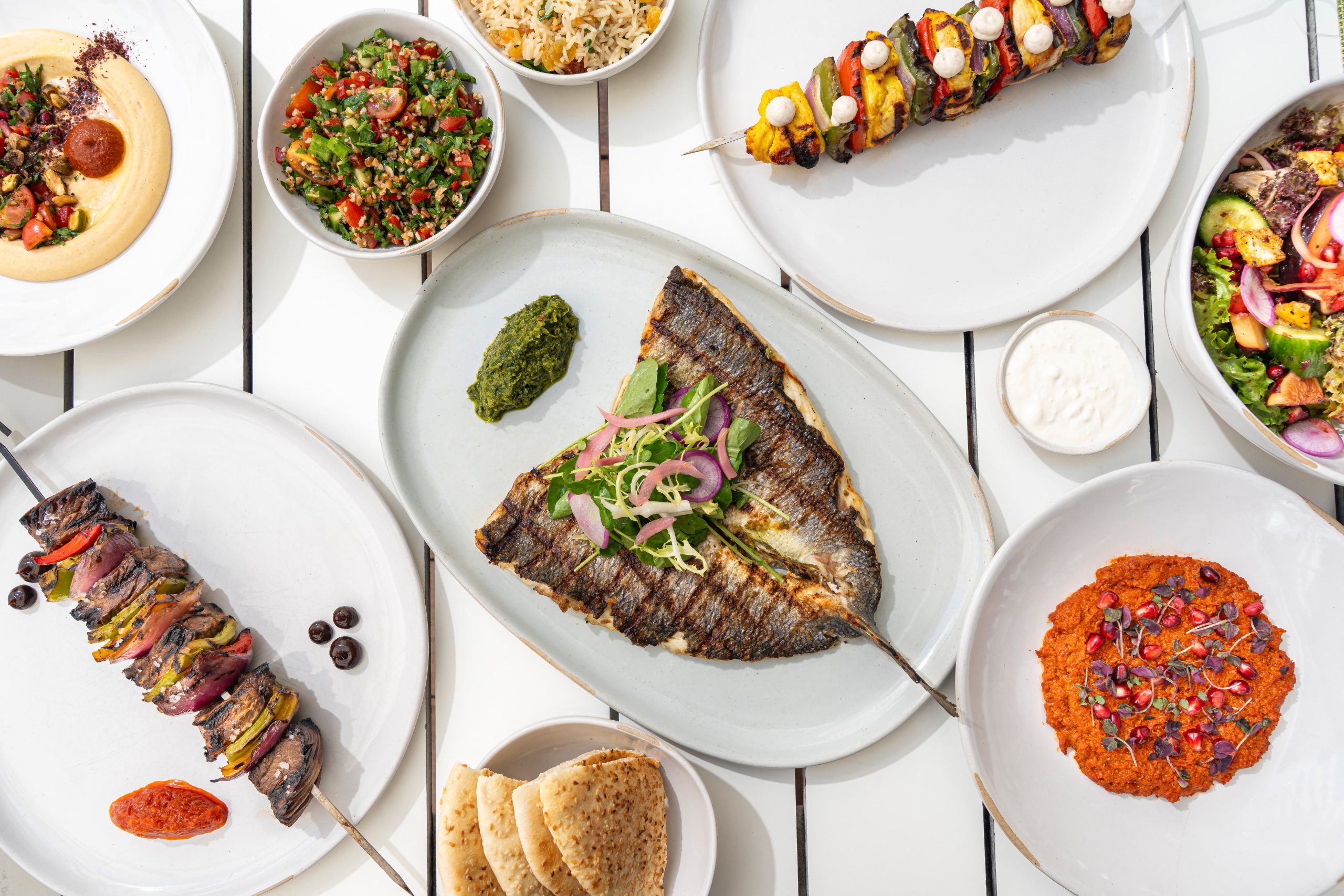 Overhead view of a Mediterranean meal spread with grilled fish, skewers, dips, salads, rice, pita bread, and sauces on a white table