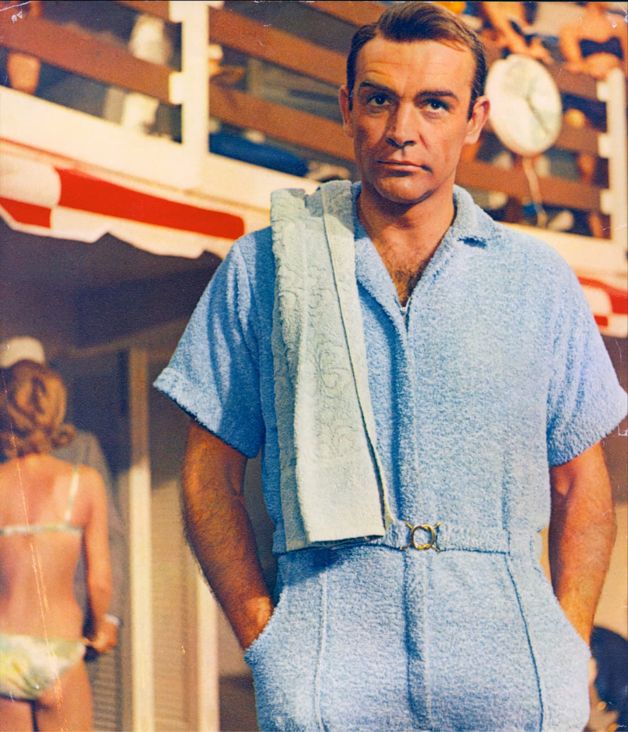 Sean Connery as James Bond wearing a light blue terry cloth playsuit with a towel over his shoulder at a poolside scene.
