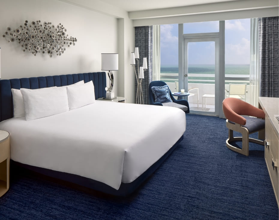 Bright guestroom with a king bed, blue furnishings, and a private balcony overlooking the ocean.