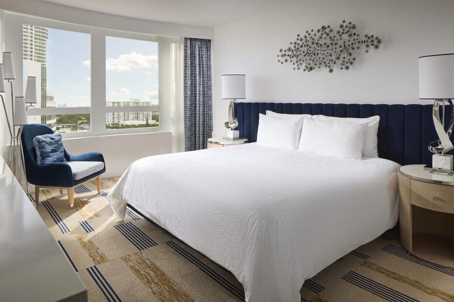 Bright junior suite bedroom with a king bed, blue accents, and large windows overlooking surrounding buildings.
