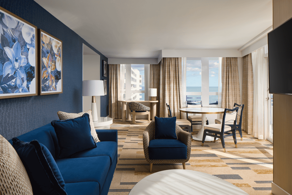 Spacious suite living area with blue seating, dining furniture, large windows, and ocean views.